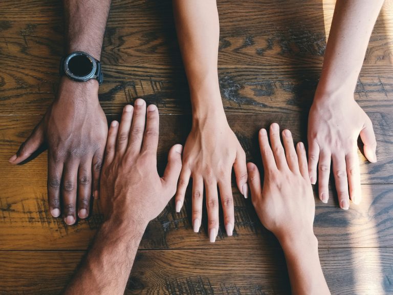 Overhead view of diverse hands—varying skin tones and genders—placed side by side on a wooden table, symbolizing intentional team composition and collaborative partnerships.