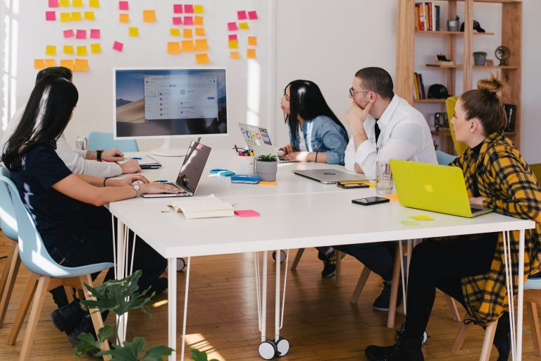 A group of people gathered around a meeting table, looking at a monitor and whiteboard covered in grouped post-it notes—representing collaborative evaluation and the importance of clear, structured scoring criteria in the RFP process.